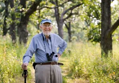 Dr. Brock standing in nature, holding a walking stick and wearing binoculars around his neck.